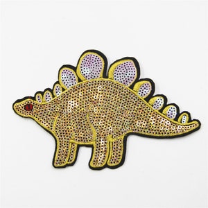 May include: A gold sequined dinosaur applique with a purple and white iridescent finish. The dinosaur has a long tail and a row of spikes down its back.