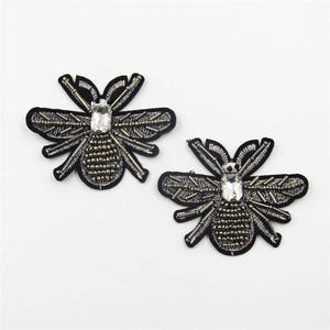 Puede incluir: Dos parches de abeja bordados en negro con detalles plateados y brillantes de cristal.