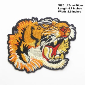 Pode incluir: Etiqueta bordada para passar a ferro com a cabeça de um tigre com a boca aberta, mostrando dentes afiados. O tigre é laranja e preto com listras brancas. A etiqueta mede 12 cm de comprimento e 10 cm de largura.