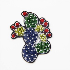 May include: A colourful embroidered cactus patch with green, yellow, blue, red and white thread. The cactus is made of felt and has a black outline.