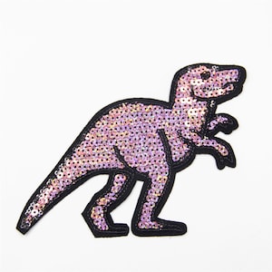 May include: A sequined dinosaur patch in shades of pink and purple, outlined in black. The patch features a Tyrannosaurus Rex design, perfect for adding to clothing or accessories. The sequins create a shimmering effect.