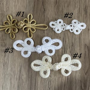 Peut inclure: Quatre fermetures décoratives en forme de grenouille, de couleur or, blanc et crème. Les fermetures sont faites de cordon tissé et présentent un motif en boucle. Chaque fermeture est numérotée de #1 à #4.
