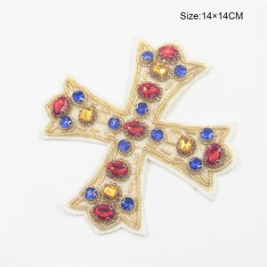 Peut inclure: Une applique en forme de croix dorée avec des strass rouges, bleus et jaunes. L'applique mesure 14 cm sur 14 cm.