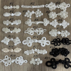 May include: A collection of decorative closures in various designs. The closures are white, cream, and black, with some featuring pearl-like beads. Styles include toggles, clasps, and decorative button closures. The closures are arranged on a wooden surface.