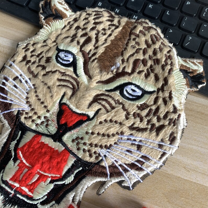 May include: Embroidered patch of a snarling leopard with brown, white, and red fur. The leopard's mouth is open, revealing sharp teeth and a red tongue.