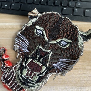 May include: A brown and black embroidered patch of a snarling tiger's face with white whiskers.