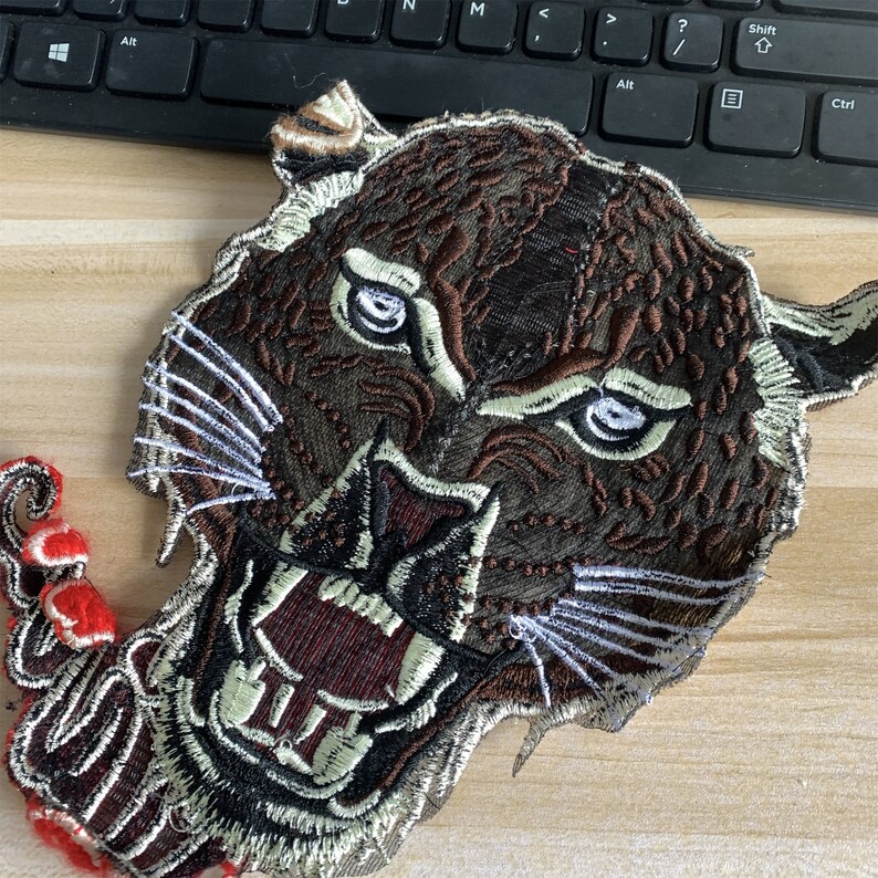 May include: A brown and black embroidered patch of a snarling tiger's face with white whiskers.