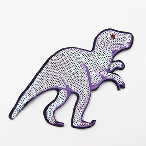 May include: Purple sequined dinosaur iron-on patch. The dinosaur is facing right and has red eyes.