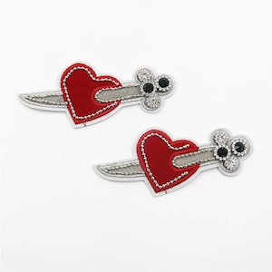 May include: Two red embroidered heart patches with a silver dagger going through them. Each heart has a silver outline and a cluster of rhinestones with green gems in the center.