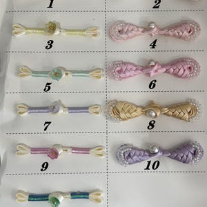 May include: A collection of eleven decorative fabric frog closures arranged in two columns. The items are numbered 1 through 11, featuring various colors including white, yellow, mint, lavender, pink, gold, and blue, with pearl and floral accents.