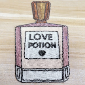 Puede incluir: Una botella de cuentas rosa y blanca con una etiqueta negra que dice "LOVE POTION" y un pequeño corazón negro.