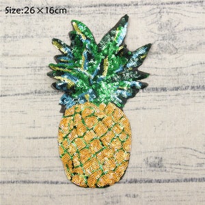 Può includere: Una toppa a forma di ananas con paillettes, di circa 26 cm per 16 cm, con corpo giallo e parte superiore verde. La toppa presenta un design dettagliato con singole paillettes, creando un aspetto strutturato e vibrante. Il testo "Size: 26 x 16cm" è visibile.