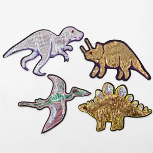 May include: Four colourful dinosaur-shaped iron-on patches with sequin details. The patches are purple, gold, and pink.