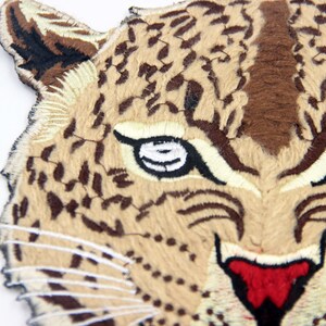 May include: Close-up of an embroidered patch featuring a brown and tan leopard face with black eyes and a red nose.