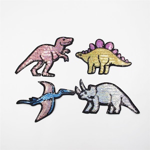 Puede incluir: Cuatro parches termoadhesivos con forma de dinosaurio con lentejuelas iridiscentes. Los parches presentan un T-Rex rosa, un estegosaurio amarillo, un pterodáctilo azul y un triceratops plateado.