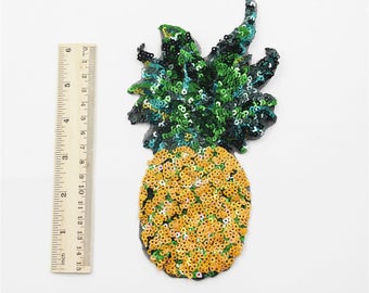 Może przedstawiać: Naszywka w kształcie ananasa z żółtych i zielonych cekinów. Ananas ma około 7,5 cm wysokości i 5 cm szerokości.