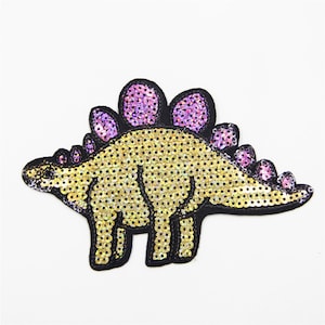 May include: A gold and purple sequined iron-on patch of a cartoon dinosaur. The dinosaur has a long tail and a row of purple spikes down its back.