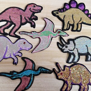 May include: A set of six colorful dinosaur and pterosaur iron-on patches. The patches are made of sequins and feature a variety of dinosaurs, including a T-Rex, a Triceratops, and a Pterodactyl.