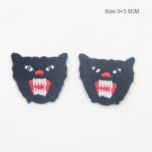 Puede incluir: Dos parches de hierro bordados negros de un rostro de gato negro gruñendo con dientes rojos y blancos. Tamaño: 3x3,5 cm.
