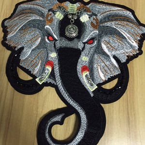 Op de afbeelding: Geborduurde strijkpatch van een grijze olifant met zwarte en witte details. De olifant heeft rode ogen en een zilveren hanger die aan zijn kop hangt.