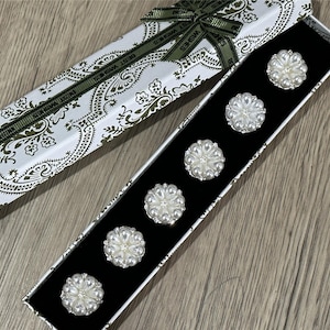 Peut inclure: Un ensemble de six boutons en forme de fleur, ornés de perles, disposés dans une boîte doublée de velours noir. La boîte blanche est décorée d'un motif cachemire et d'un ruban vert avec l'inscription "BEST WISHES".