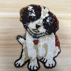 Puede incluir: Un parche de lentejuelas con la forma de un perro San Bernardo. El parche presenta un cuerpo blanco con marcas marrones y negras, un collar azul y una etiqueta roja en forma de corazón. El perro está sentado con una pata levantada.