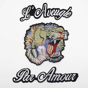 Puede incluir: Tres parches bordados para planchar. El parche superior es negro con texto blanco que dice "L'Aveugle". El parche del medio es una cabeza de tigre colorida con un contorno negro. El parche inferior es negro con texto blanco que dice "Par Amour".