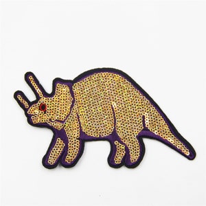 May include: A gold sequined iron-on patch of a dinosaur with purple trim. The dinosaur has a red eye.