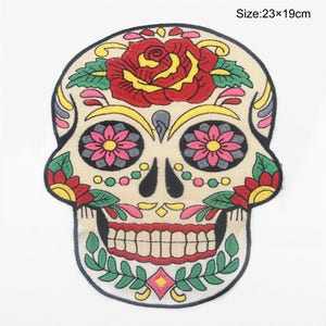 Könnte beinhalten: Gestickter Zucker Schädel Patch mit einer roten Rose, rosa und gelben Blumen und grünen Blättern. Der Schädel ist beige mit schwarzen Umrissen und einem roten Lächeln. Größe: 23x19cm.