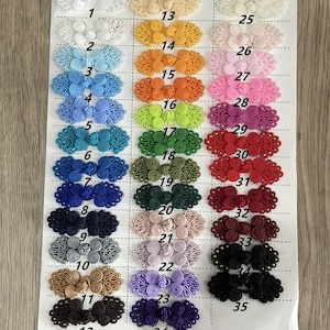 May include: A collection of thirty-five decorative frog closures arranged in three columns on a white sheet. Each closure features a symmetrical, coiled cord design in various solid colors, including white, yellow, orange, green, blue, red, and black, with each item numbered from 1 to 35.
