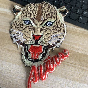 May include: Embroidered iron-on patch featuring a snarling leopard with a red mouth and white teeth. The word "Alive" is embroidered in red and white below the leopard's head.