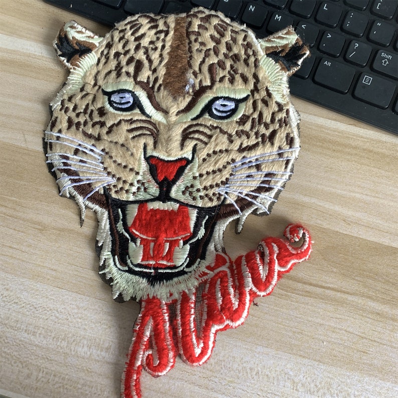 May include: Embroidered iron-on patch featuring a snarling leopard with a red mouth and white teeth. The word "Alive" is embroidered in red and white below the leopard's head.