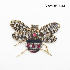 Puede incluir: Una aplicación de abeja de cuentas doradas y negras con diamantes de imitación rojos y transparentes. La abeja mide 7 x 10 cm.