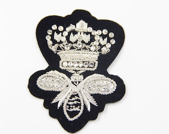 May include: A black velvet patch featuring a silver embroidered crown and bee design. The crown and bee are embellished with clear rhinestones.