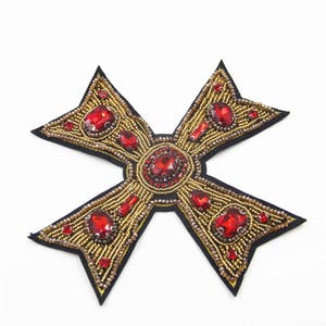May include: A gold and red cross-shaped patch with a beaded design and red rhinestone accents.