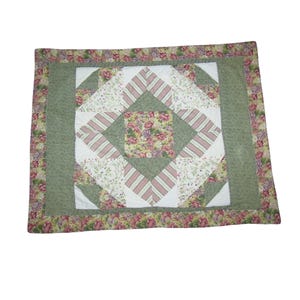 May include: A square quilt with a floral pattern. The center features a diamond shape with a floral print, surrounded by white and pink striped squares. The border is a solid green, with a floral patterned edge.