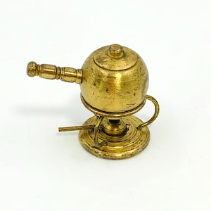 May include: A small, antique brass kettle with a round body, a lid, and a handle. The kettle sits on a circular base with a small, curved handle. The brass has a warm, aged patina, suggesting its vintage.