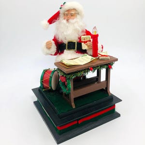 May include: A Christmas-themed figurine of Santa Claus, dressed in a red suit with a black belt, holding letters. A small wooden desk with a candle and a drum are also present. The base is decorated with green and red accents.