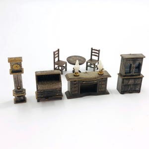 May include: A miniature furniture set featuring a tall grandfather clock, a drop-front desk, a round table with two chairs, a wide desk with two small white lamps, and a hutch cabinet. All pieces are finished in a dark, weathered metallic tone.