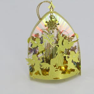 May include: Gold-colored Christmas ornament with an arched shape, featuring angel silhouettes and a snowflake design. The ornament has a clear, iridescent background and a gold hanging loop. A festive holiday decoration.