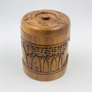 May include: A cylindrical wooden box with a lid. The box is a medium brown color and features intricate carved designs around the body and on the lid. The lid has a small, round knob in the center.