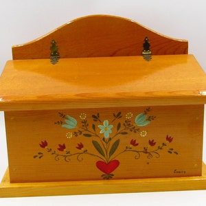 May include: A light brown wooden wall-mounted box with a hinged lid and a curved back panel. The front features a hand-painted floral design with blue and red flowers, green stems, and a red heart at the base. The name Eunice is painted in the corner.