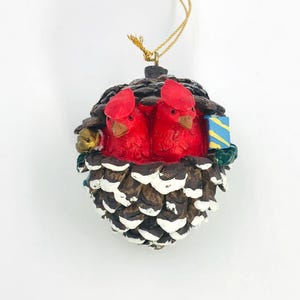 May include: A Christmas ornament shaped like a pine cone, featuring two red cardinal birds. The ornament has a gold string for hanging and includes a small bell and a blue and yellow candle.