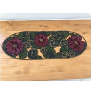 May include: An oval-shaped table runner with a beaded design. The runner features three large, burgundy poinsettia flowers with sequin centers, surrounded by green leaves and swirling accents. The background is dark green with gold-colored beads.