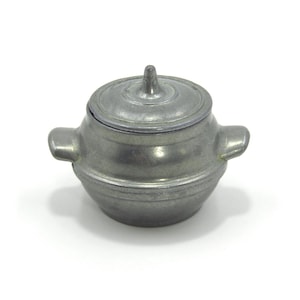 May include: A small, antique-style metal pot with a lid. The pot is a dull silver color and has two handles on the sides. The lid has a small knob on top. The pot is likely used for cooking or storage.