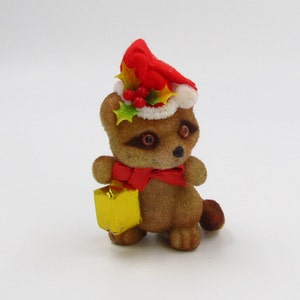 May include: A plush raccoon wearing a red Santa hat with green holly and red berries. The raccoon is holding a gold gift box and has a red scarf around its neck.