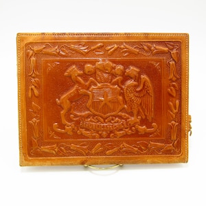 May include: A brown leather embossed clutch with a detailed design of the coat of arms of Chile. The design includes a star, a shield, a condor, and a llama. The text "REPUBLICA DE CHILE" is embossed below the design.