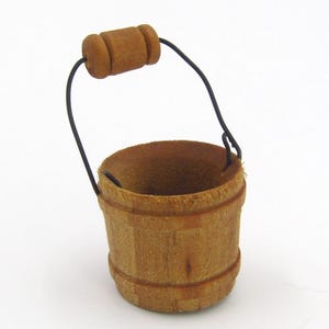 May include: A small, wooden bucket with a dark wire handle and a wooden handle grip. The bucket is light brown with darker brown bands around the top and bottom. The bucket is empty and sits on a white background.