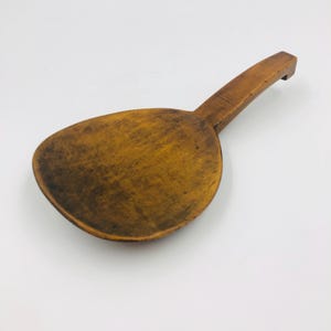 May include: A wooden spoon with a wide, oval bowl and a long, flat handle. The spoon is made of a light brown wood with visible grain and a slightly worn appearance. The handle has a small notch at the end.