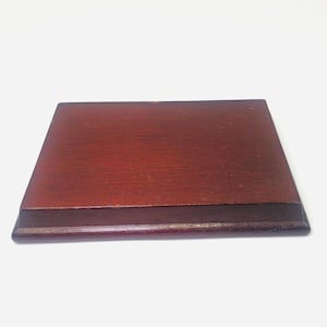 May include: A rectangular wooden display base with a dark reddish-brown finish and a smooth, flat top surface. The base features beveled edges that slope downward toward the bottom, creating a tiered profile with a polished, wood-grain texture.